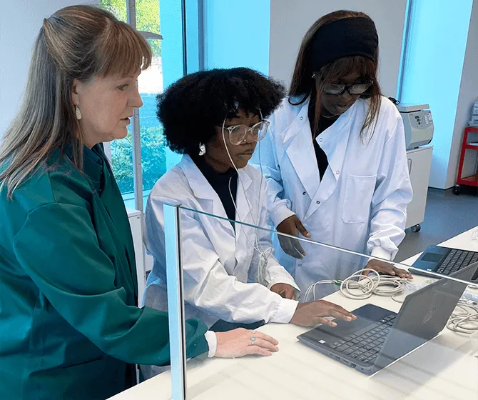 Dr. Ann Rajnicek is pictured in the lab helping two students in white lab coats to perform EEG using a laptop.