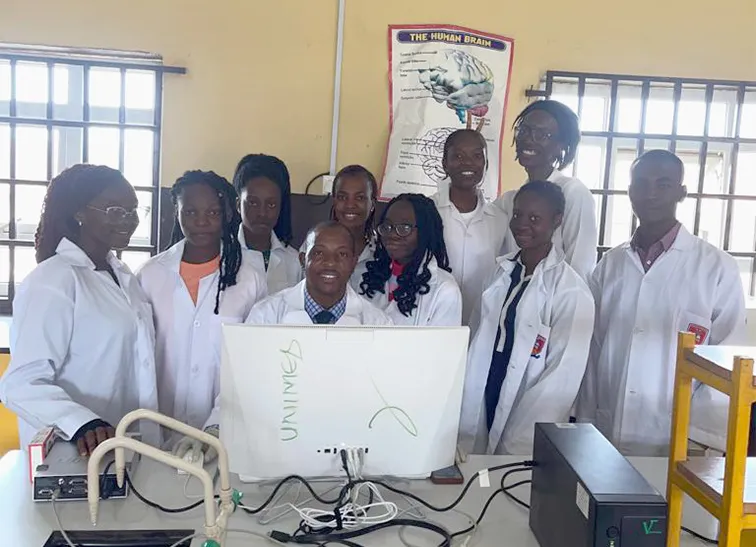 Johnson Olowe sits behind a desktop computer screen. He is surrounded by 9 students, who stand around him in a laboratory environment.