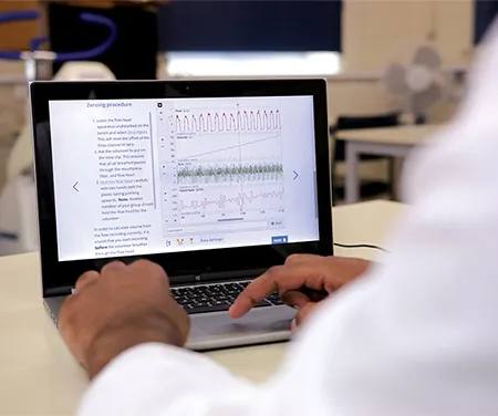 A photo of a laptop on a table. There is data on the laptop screen. The arm of someone wearing a white lab coat is in the foreground of the shot, and their hands rest on the laptop trackpad.