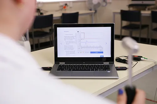 A photograph over the shoulder of a student wearing a white lab coat in the lab. The student is holding a tendon hammer which is blurred in the foreground of the image. The center of the image is taken up by a laptop showing example data in Lt.