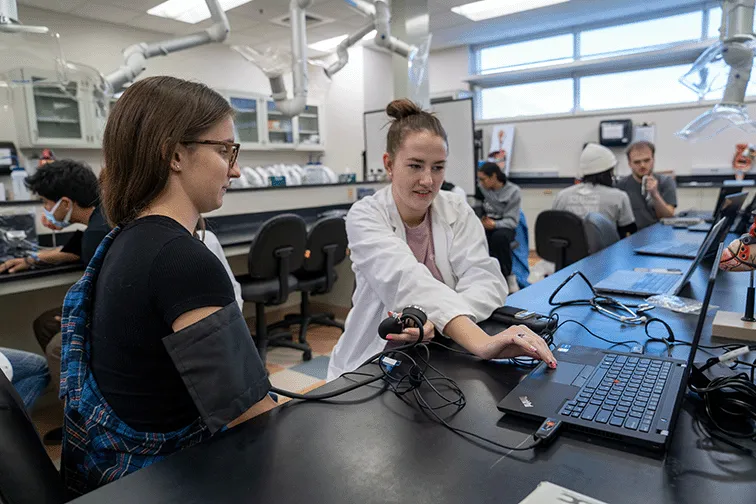 A photograph of two students in a lab space, with one measuring the blood pressure of the other with a blood pressure cuff connected to a laptop.