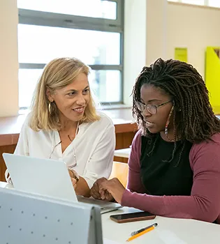 A photograph of a professor with a student. The student is looking at a laptop and the professor is looking at the student and smiling.