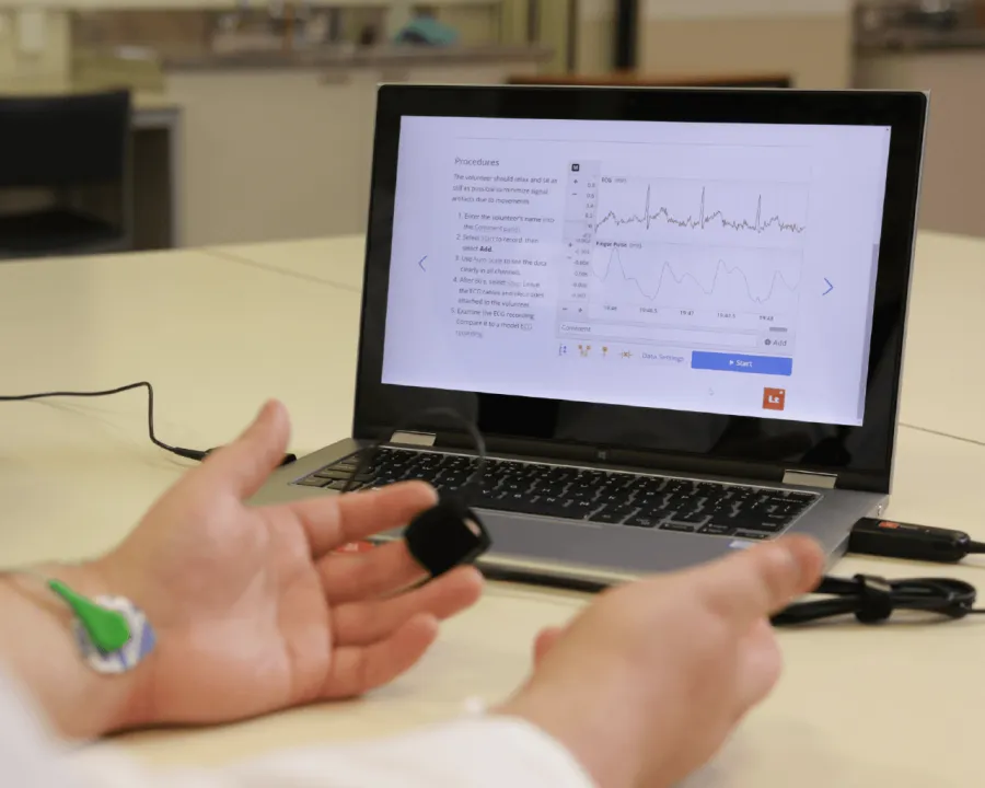 A photograph of a laptop on a table in a physiology lab environment. The software platform Lt is shown on screen, with example physiological data in the data sampling panel. A person's hands are resting in the foreground in front of the laptop, connected to a pulse transducer and a biopotential sensor (via electrodes).