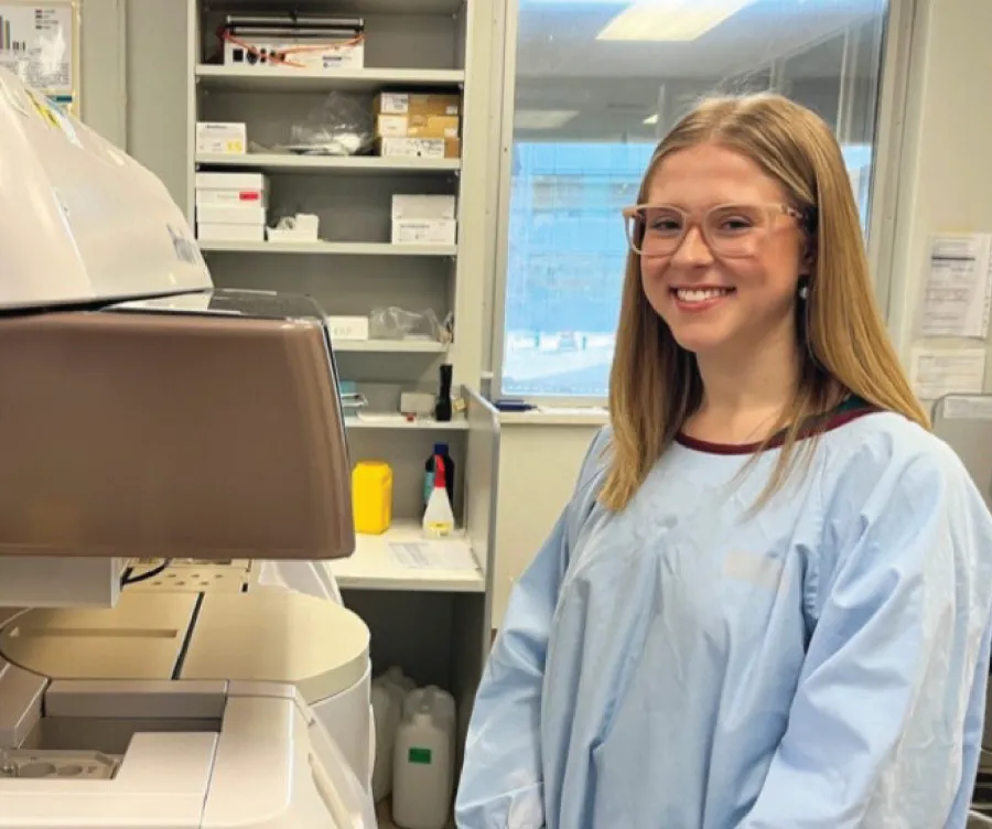 A photograph of Hannah Gapps smiling in a lab setting and wearing light blue scrubs.