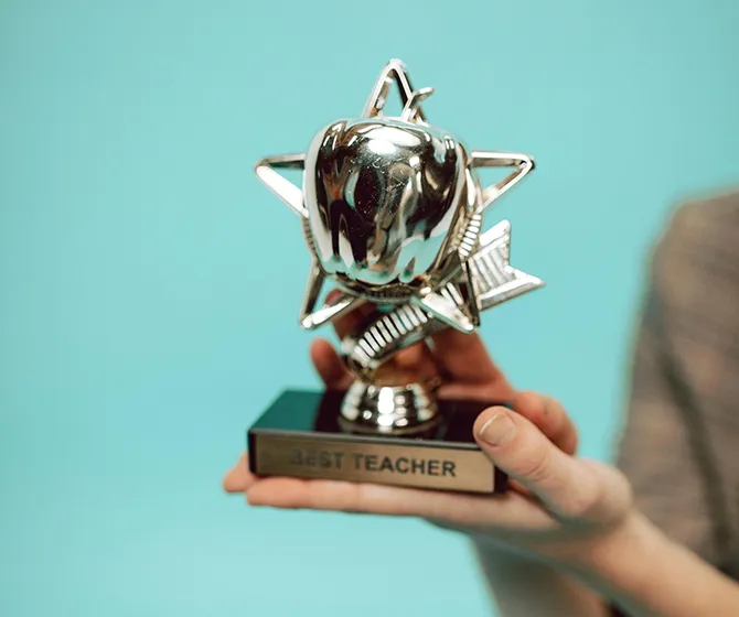 A photograph of a pair of human hands holding a silver trophy of an apple overlaid on a star. The trophy reads, "BEST TEACHER".